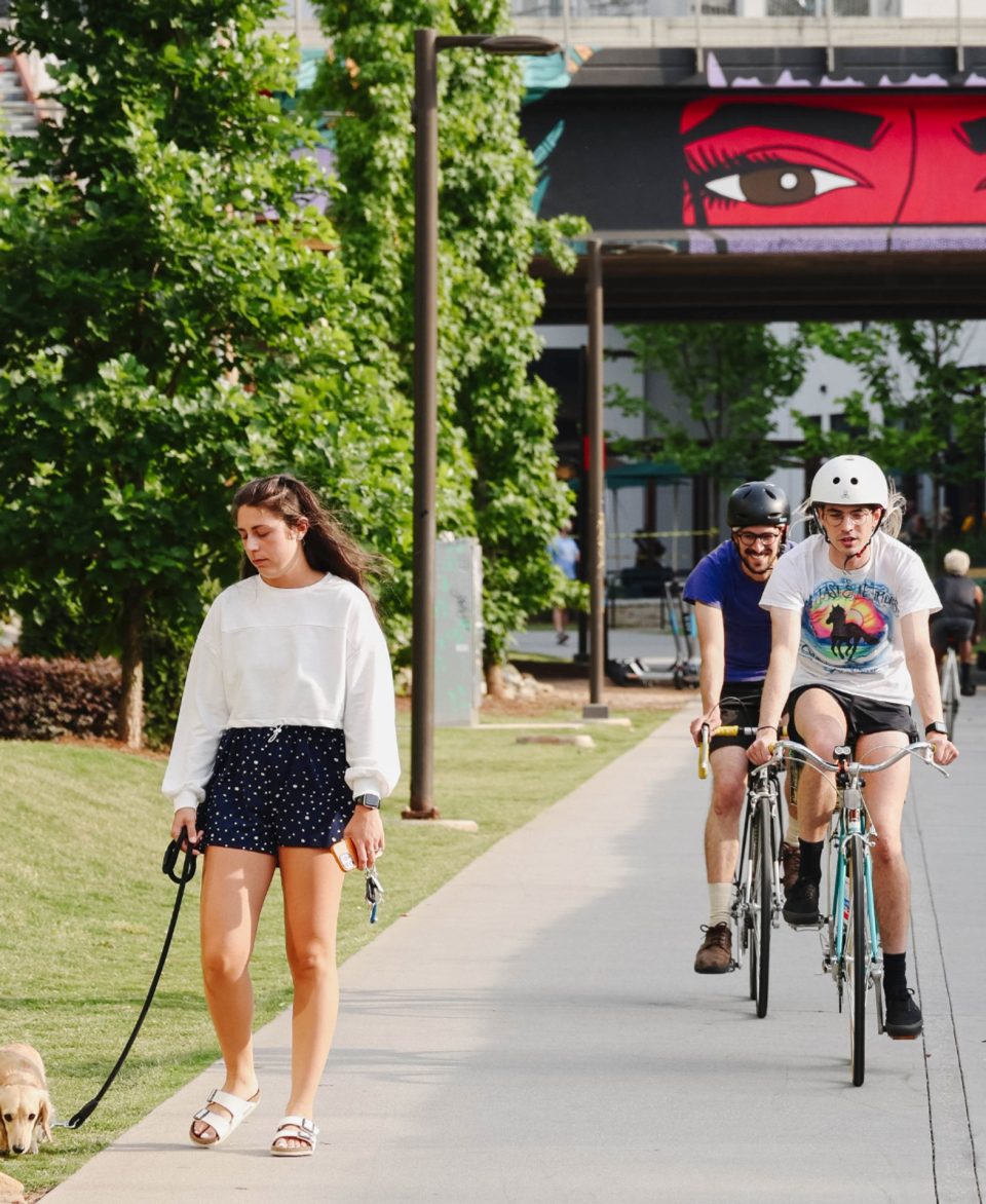Pedestrians & cyclists on a paved trail lined with trees & public art